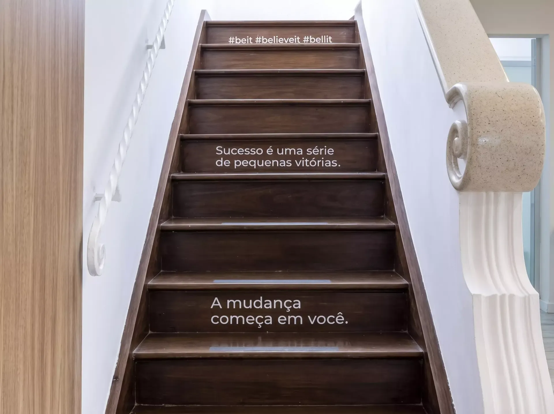 Escadaria com paredes brancas e degraus escuros, com texto em português em branco nos espelhos dos degraus.