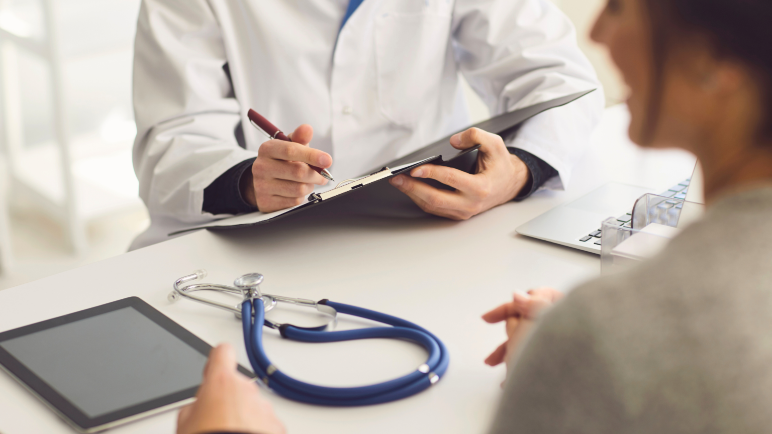 Doctor writing notes during a consultation, with a blue stethoscope on the desk and a tablet in view