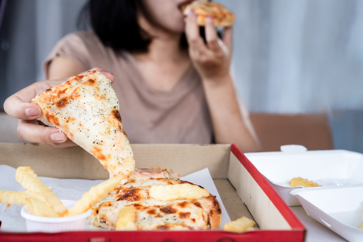 Person lifting a slice of pizza from a box, with more pizza and fries on the table.