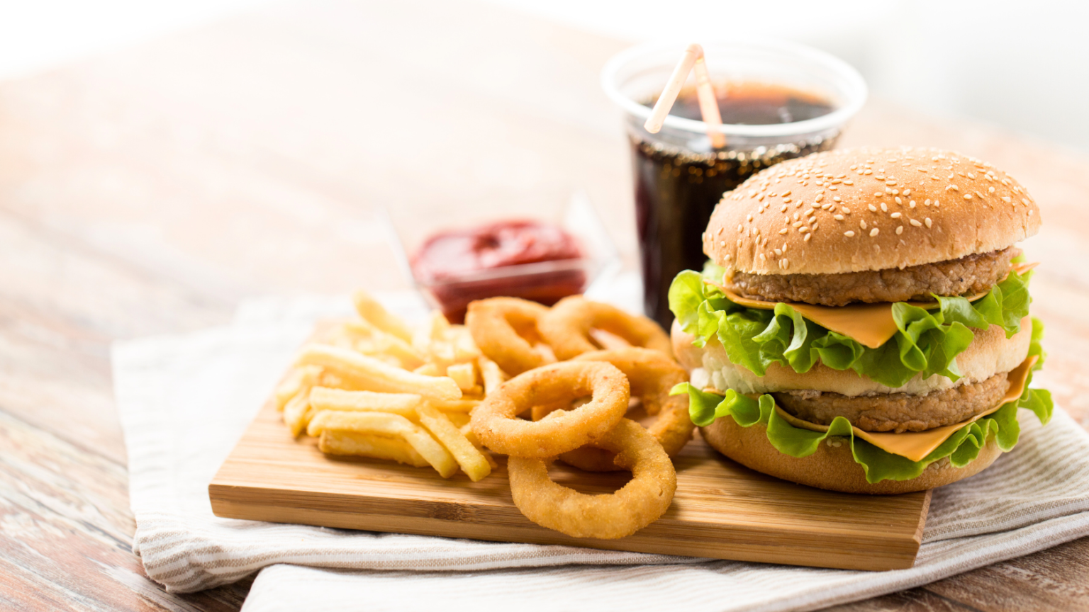 Burger with fries and onion rings on a wooden board, with a drink in the background