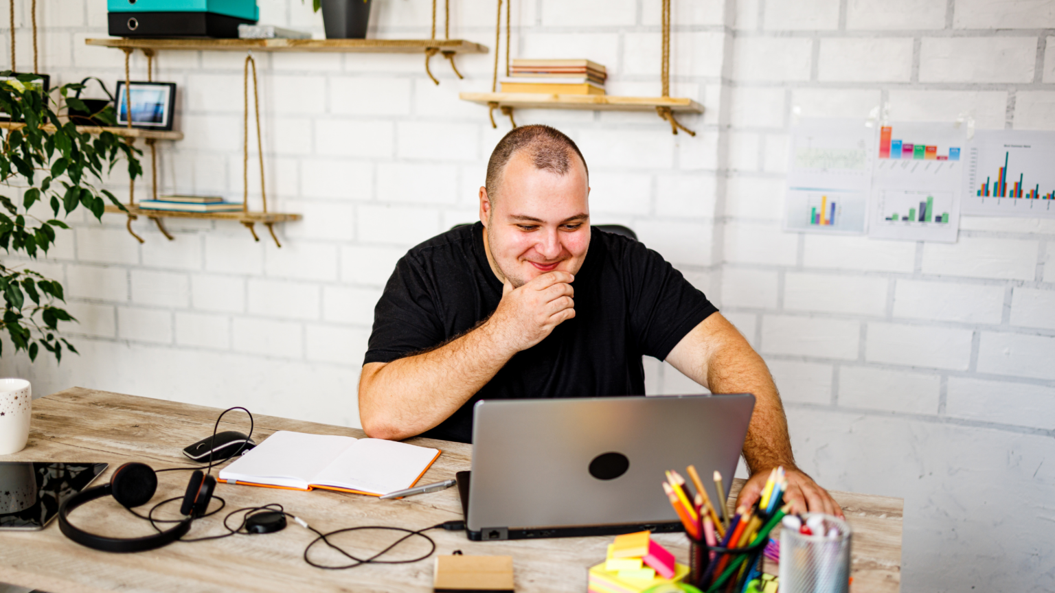 Man in black shirt working at a laptop in a bright kitchen, hand over mouth, with headphones and stationery on the table