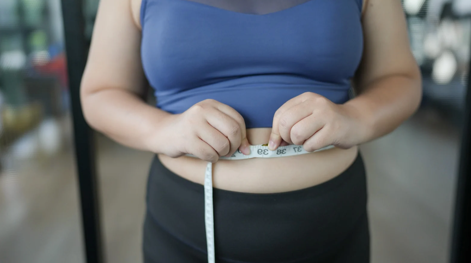 Person measuring waist with a tape measure, wearing a blue top and black bottoms.
