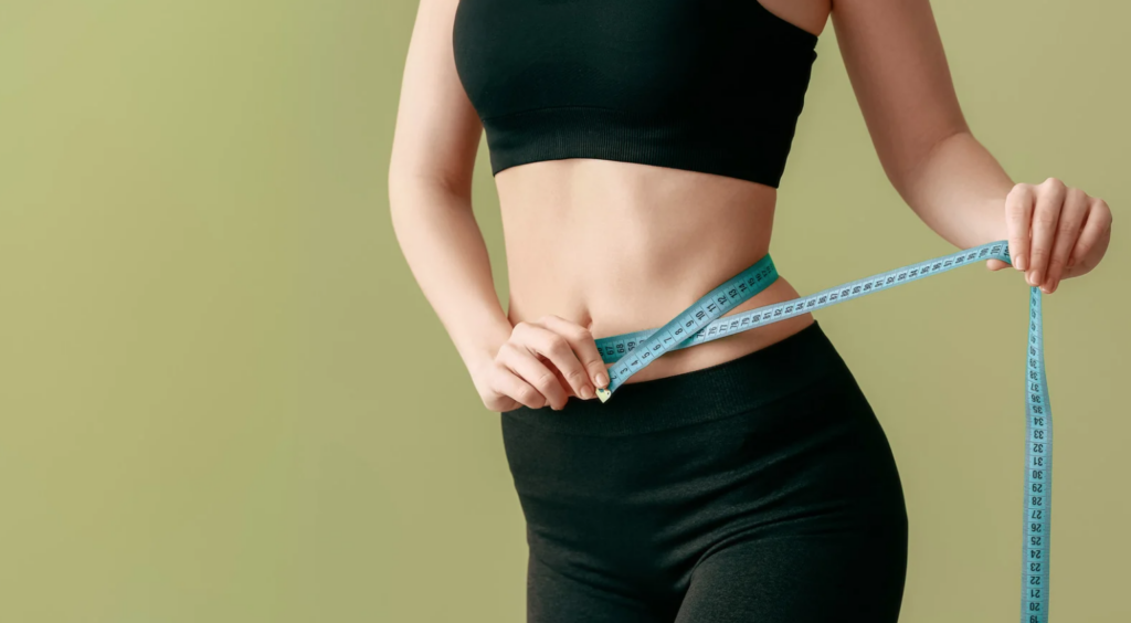 Person measuring their waist with a blue tape measure, wearing black athletic clothes, on a plain green background