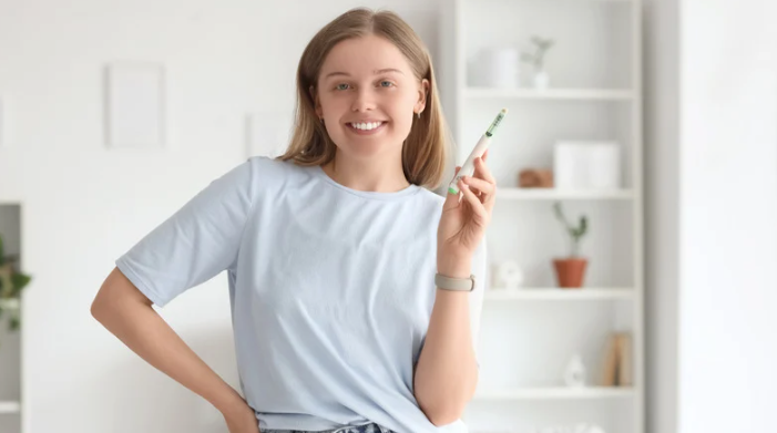 Pessoa sorridente de camisa azul-clara segurando uma caneta em uma sala iluminada com prateleiras 