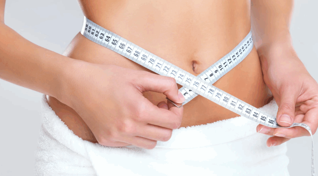 Mãos medindo a cintura com uma fita métrica branca sobre a barriga descoberta.
