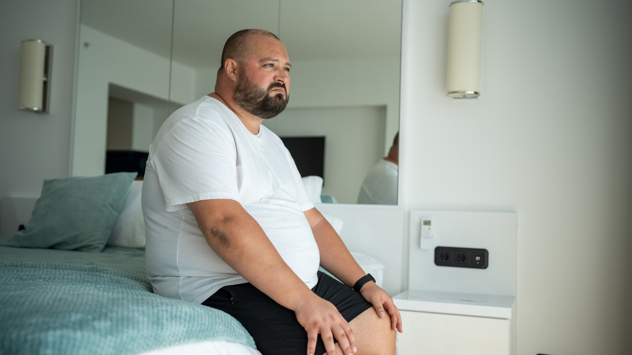 Man sitting on a bed in a bright bedroom, looking out of frame.