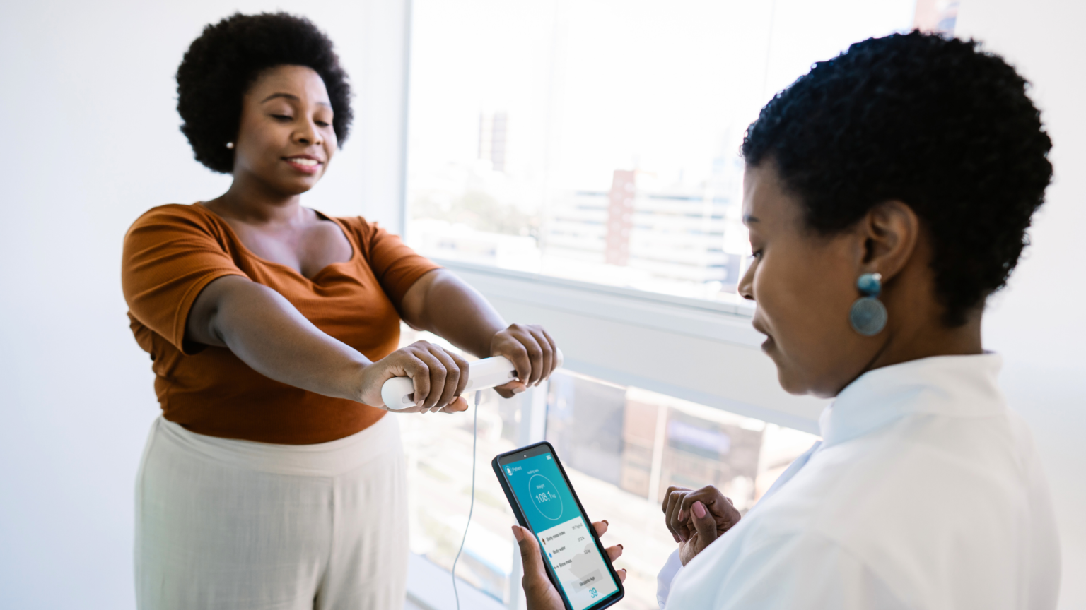 Two women at a counter, one holding a smartphone and the other holding a bottle, in a bright room.