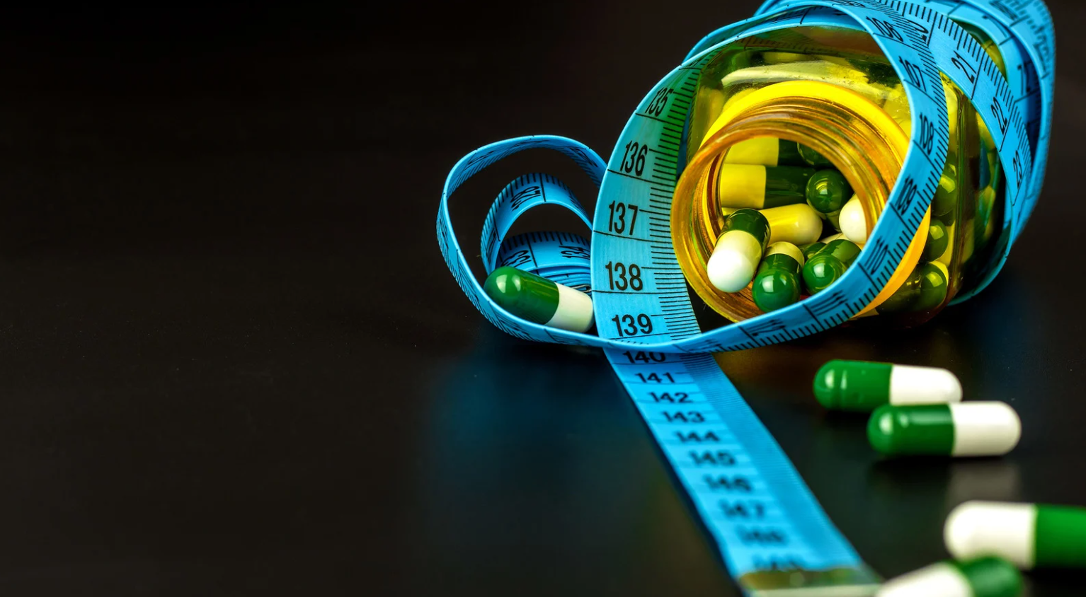 Blue measuring tape wrapped around yellow capsules and green pills on a black background