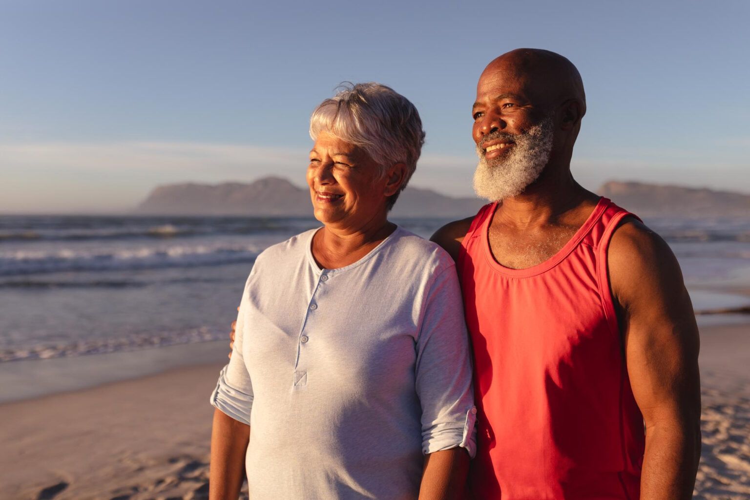 Two people standing on a beach at sunset, smiling and looking out at the water