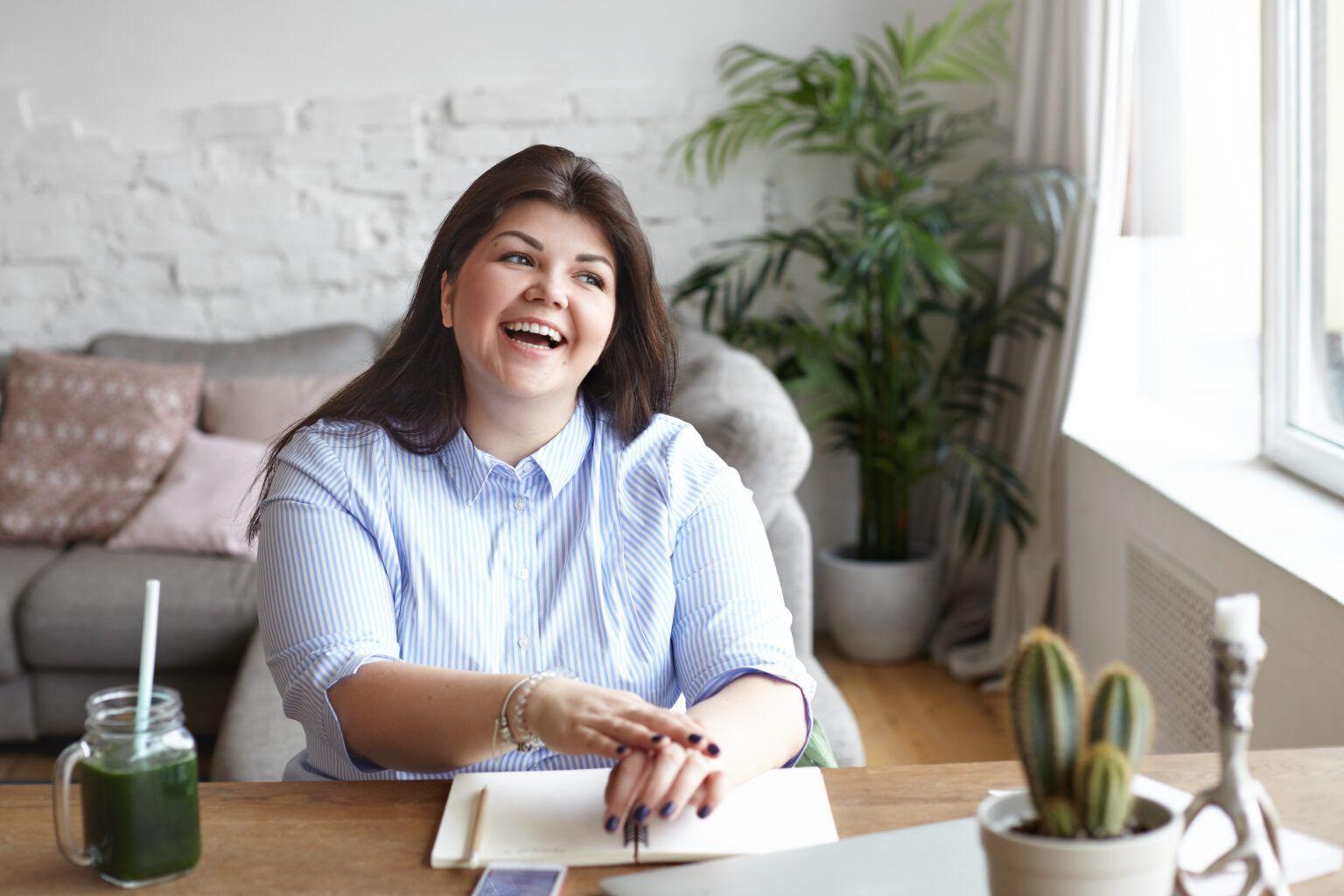 Smiling person writing at a desk in a bright home office with plants and a window