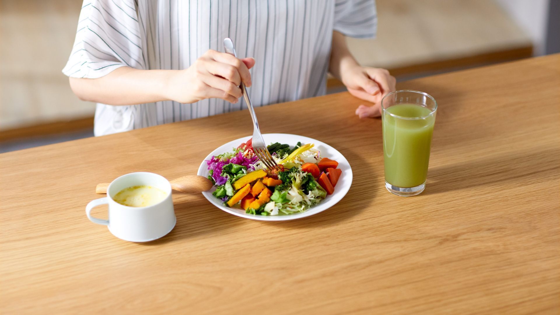 Person eating a colorful salad with soup and a green drink on a wooden table