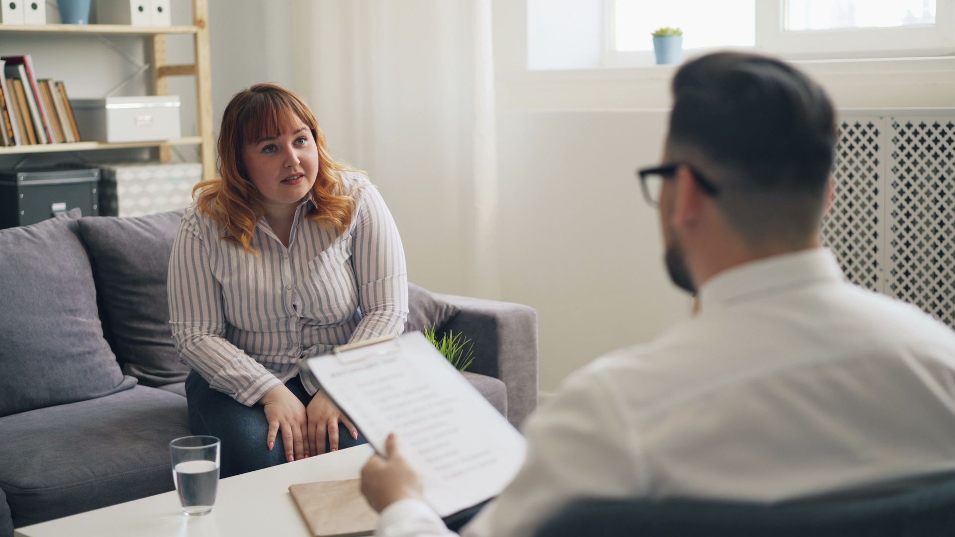 Therapist speaking with a seated client in a bright living room, holding notes beside a couch.
