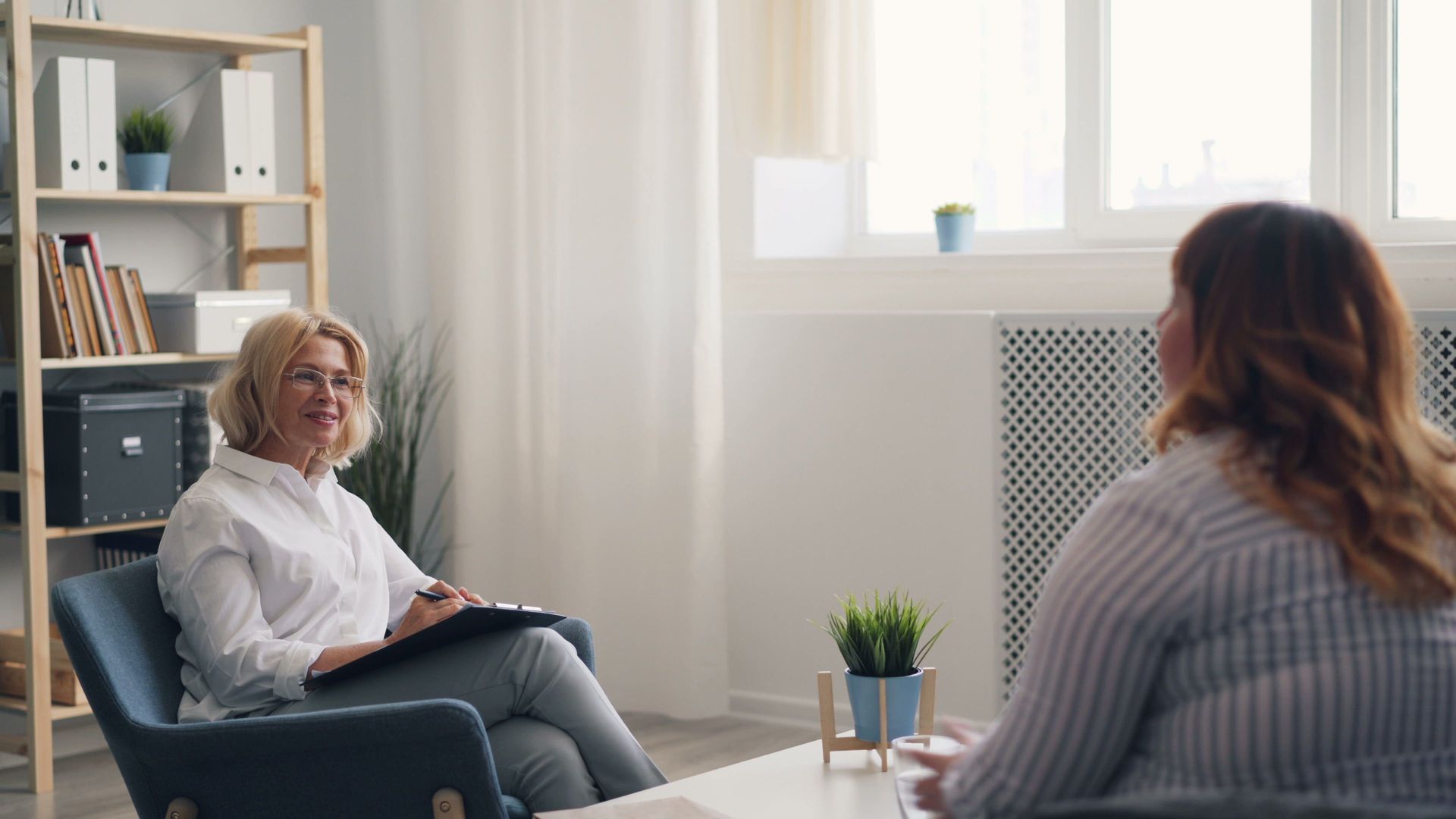 Therapist and client talking in a bright office with a plant and bookshelf.
