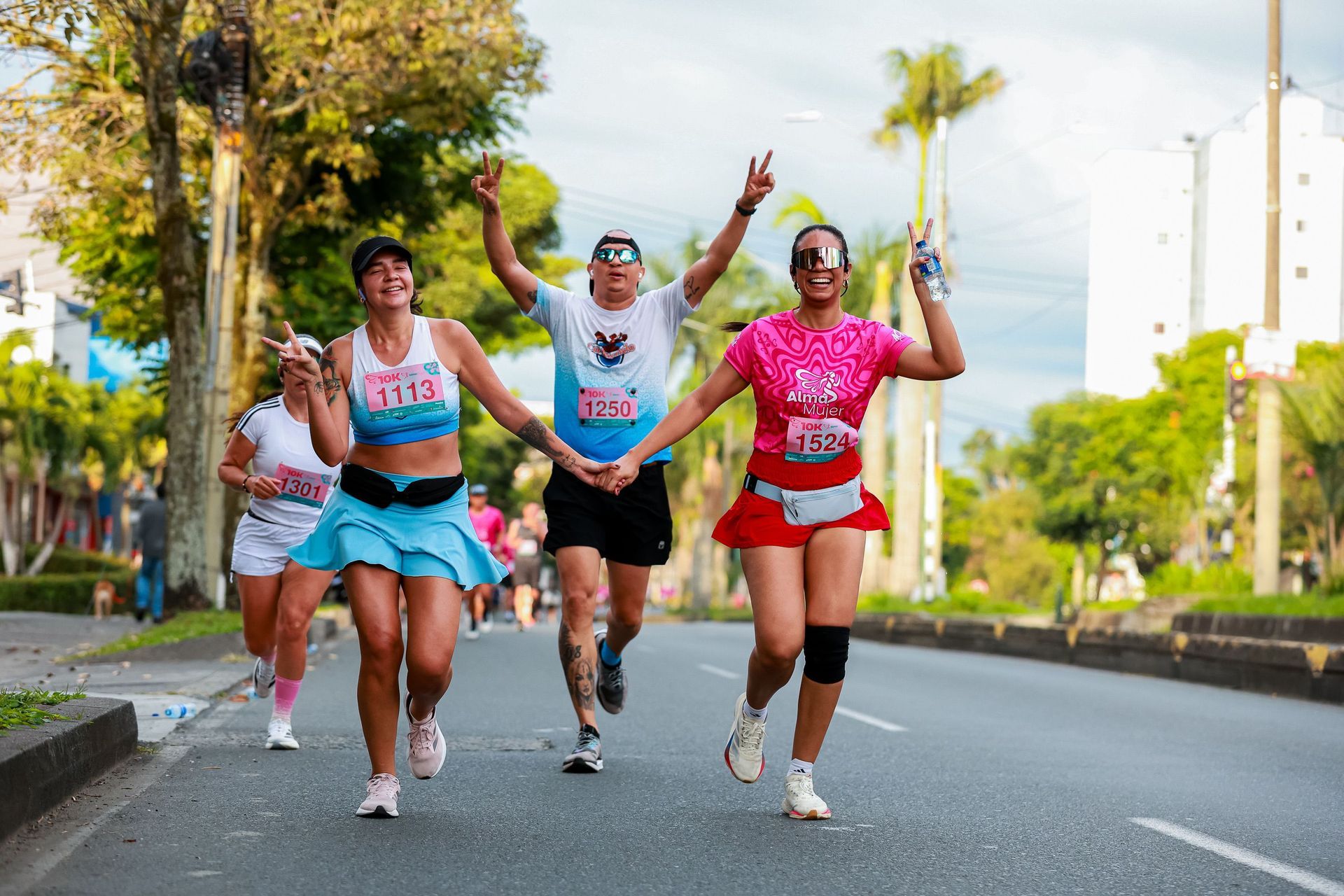 Three runners in colorful shirts celebrate while jogging on a sunny palm-lined road.