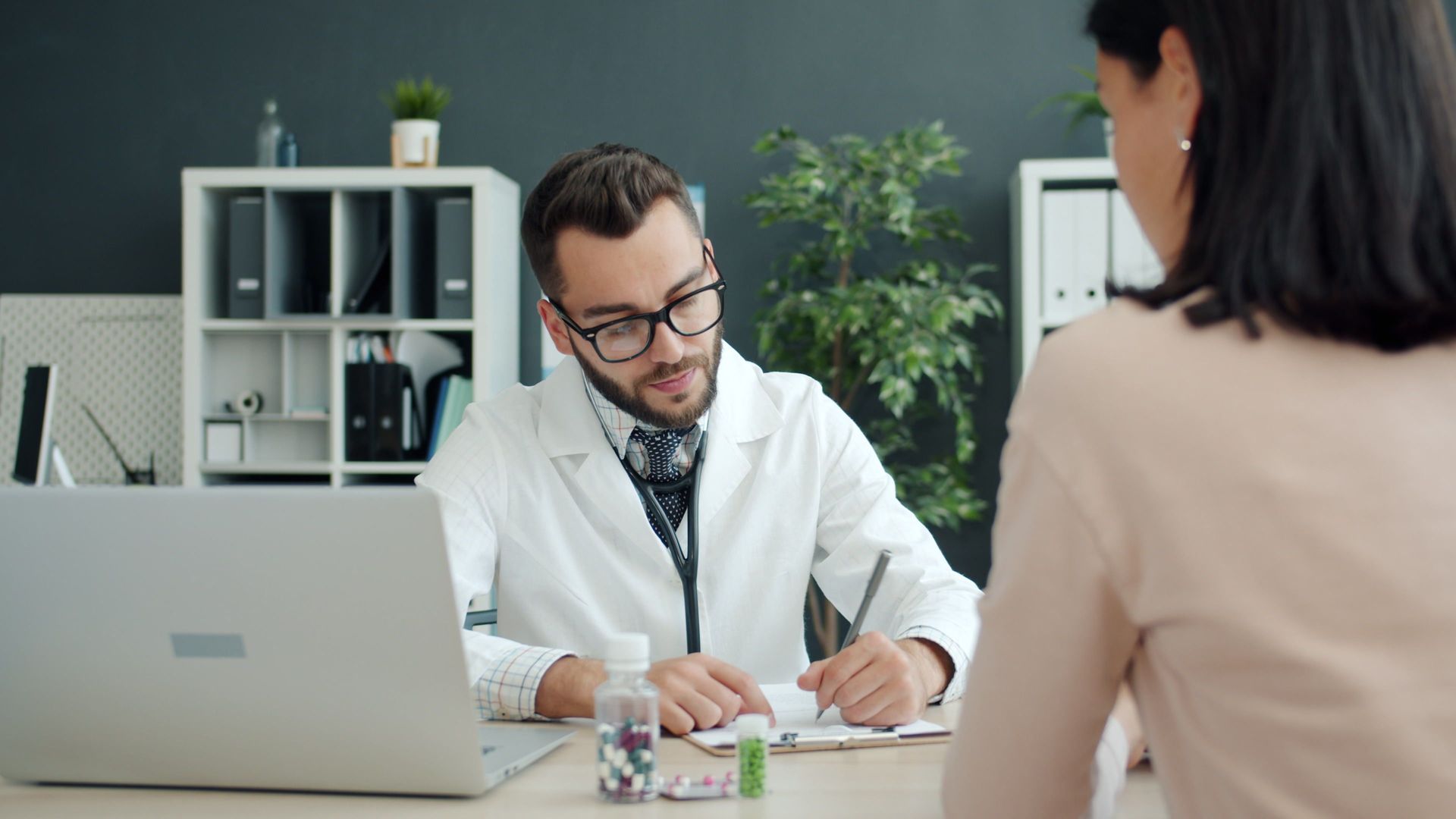 Doctor consulting a patient across a desk in a bright clinic office, with a laptop and medical samples nearby