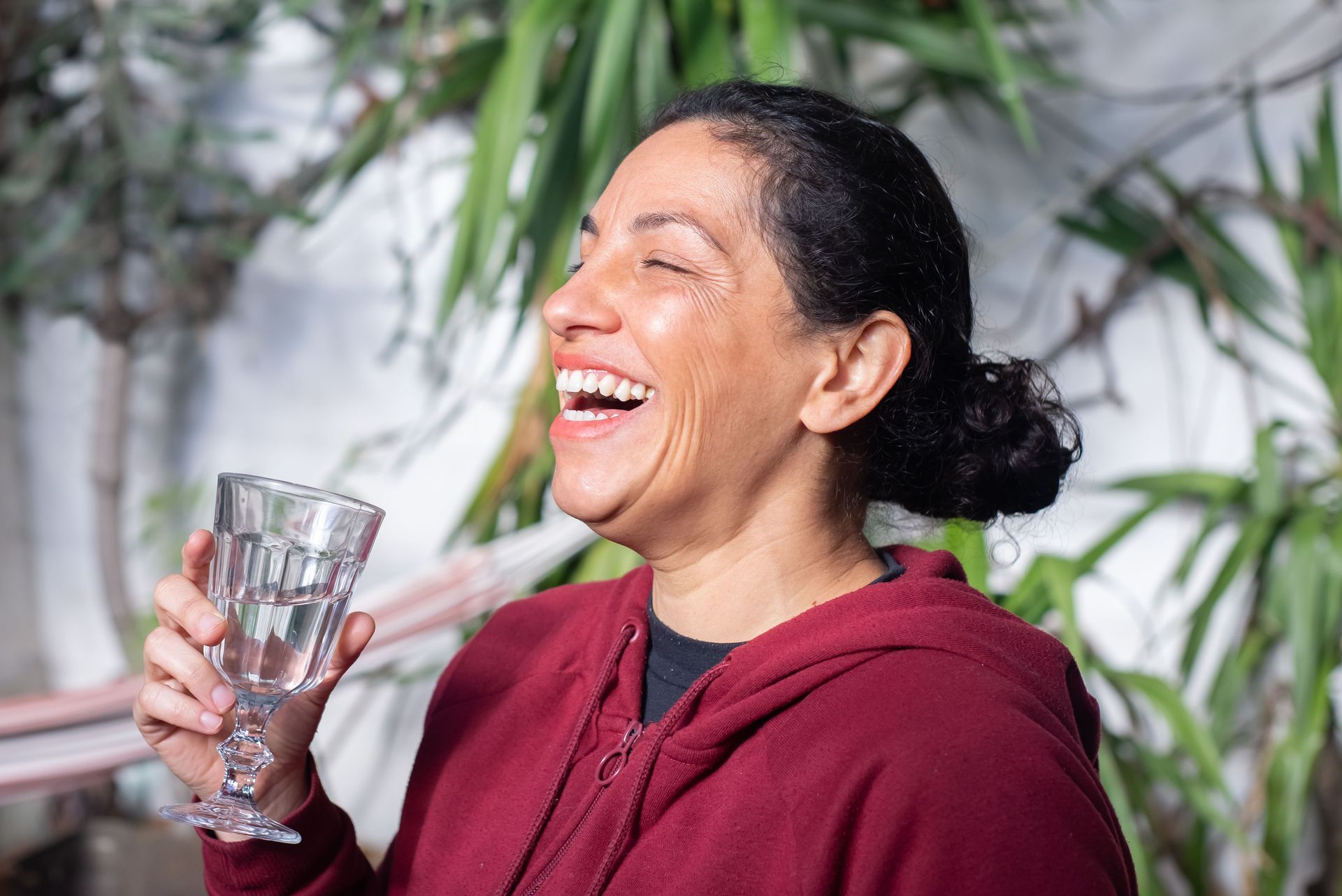 Woman in a maroon hoodie laughing while holding a glass in a leafy outdoor setting