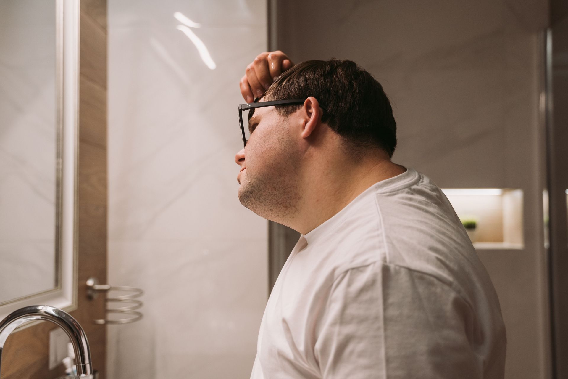 Man brushing teeth in a bathroom mirror, wearing a white shirt and glasses.