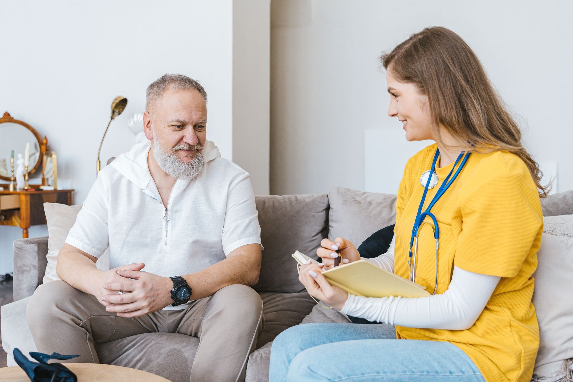 Two people talking on a couch, one holding a notepad in a bright living room.