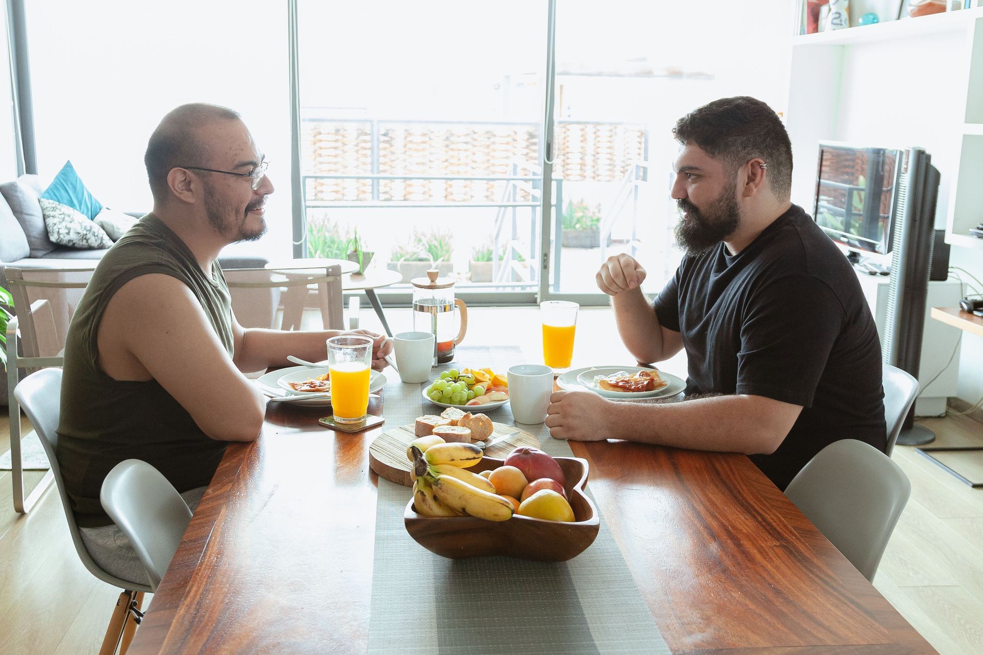 Two people talking at a dining table with drinks and fruit in a bright kitchen
