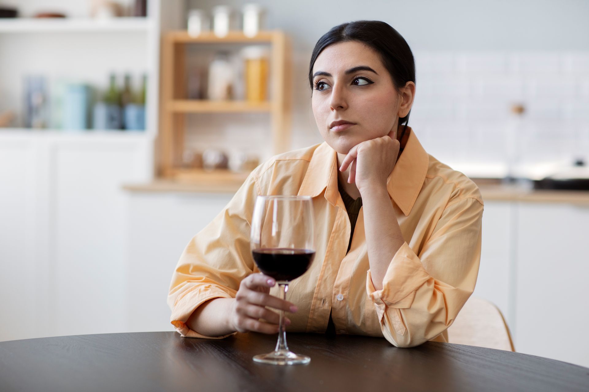 Woman resting at a table, holding a glass of red wine and gazing thoughtfully in a bright kitchen