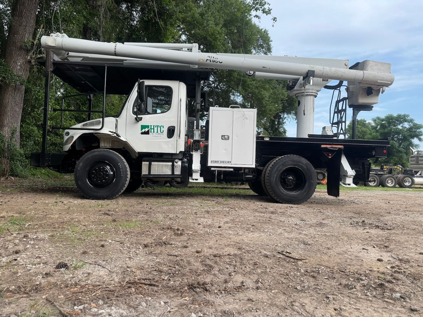 White utility truck with mounted boom parked on a dirt lot beside trees and equipment.