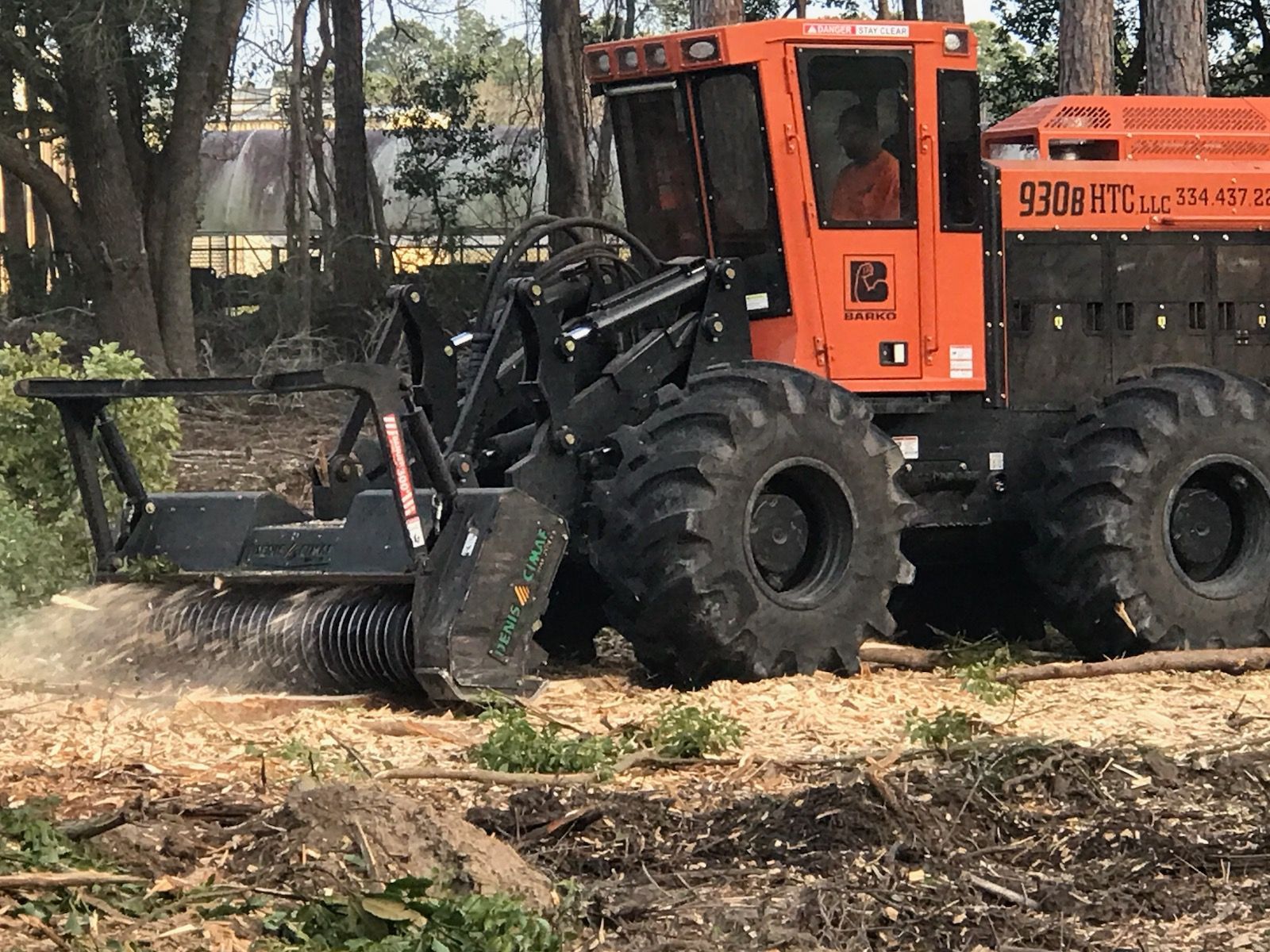 Orange forestry mulcher clearing brush in a wooded area, kicking up dust.