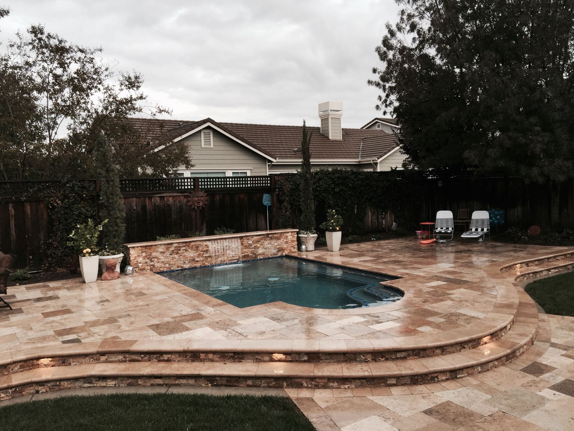 Backyard pool with stone patio, water feature, two lounge chairs, and cloudy sky.