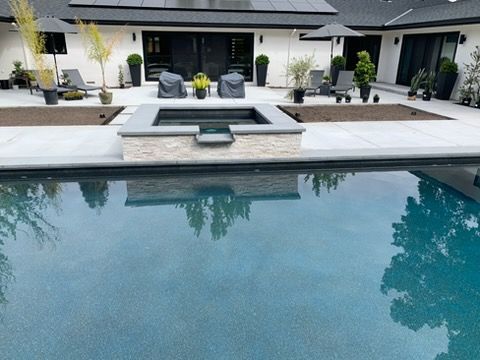 Pool and spa in a backyard patio. Stone patio with lounge chairs, trees, and dark trim.