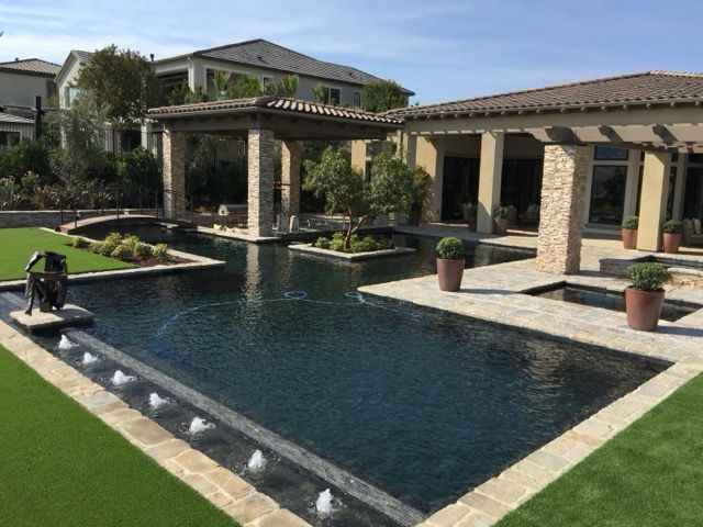 Luxurious backyard with a dark blue pool, fountains, and stone patio under a pergola, adjacent to a house.