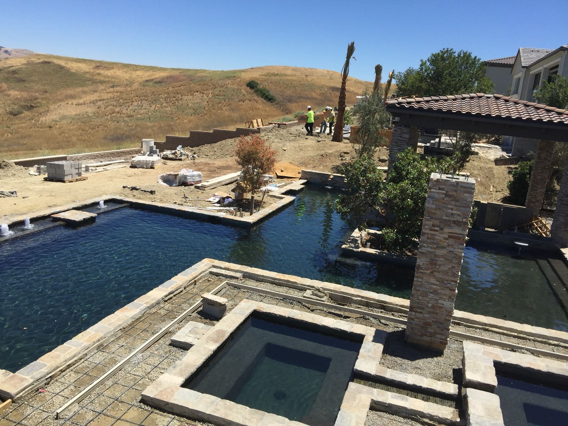 Pool and spa construction site with stonework, water, and hillside in the background.
