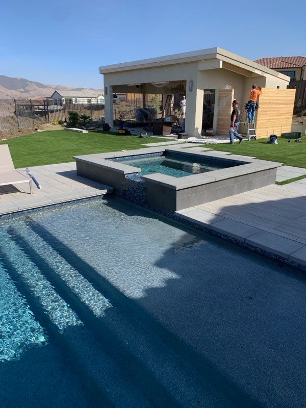 Pool and spa with adjacent cabana on a sunny day. Person working on the cabana, green grass, and light blue water.