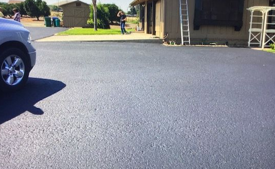 A gray asphalt driveway with a car parked on the left. A ladder leans against a brown building.