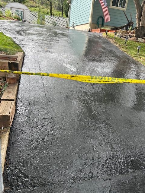 Wet, freshly sealed asphalt driveway blocked by yellow caution tape, in front of a blue house with an American flag.