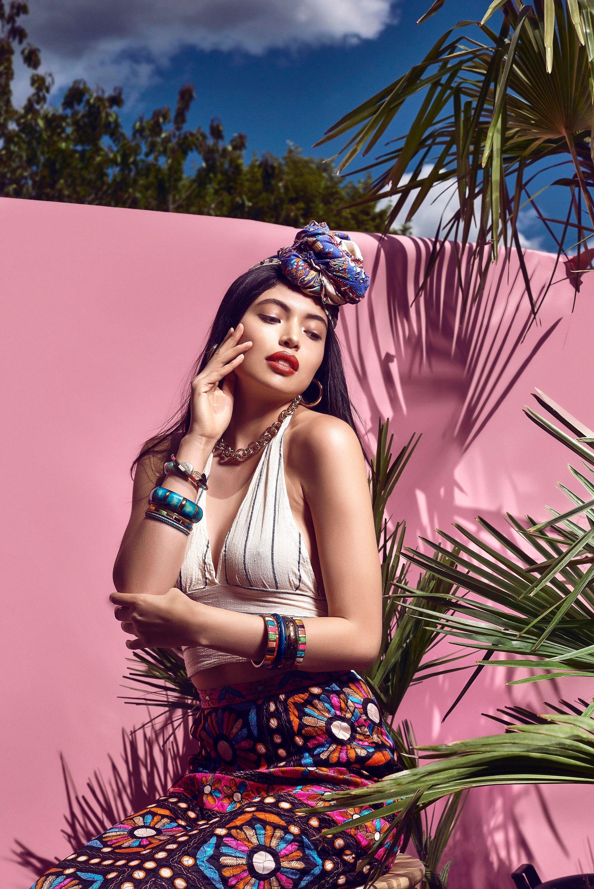 A woman is sitting in front of a pink wall next to a palm tree.