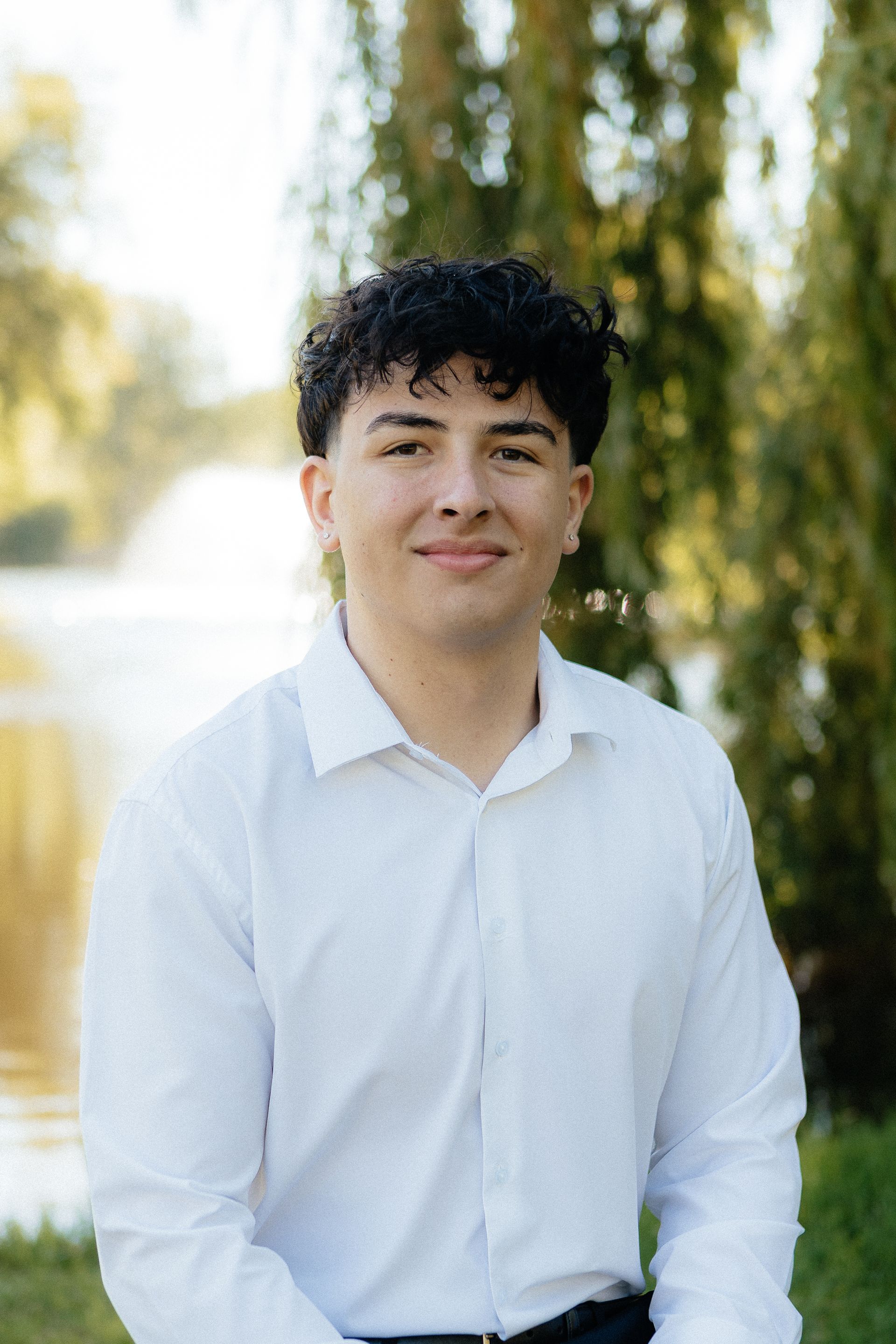 Person in a white shirt standing outdoors by a sunlit fountain and trees.