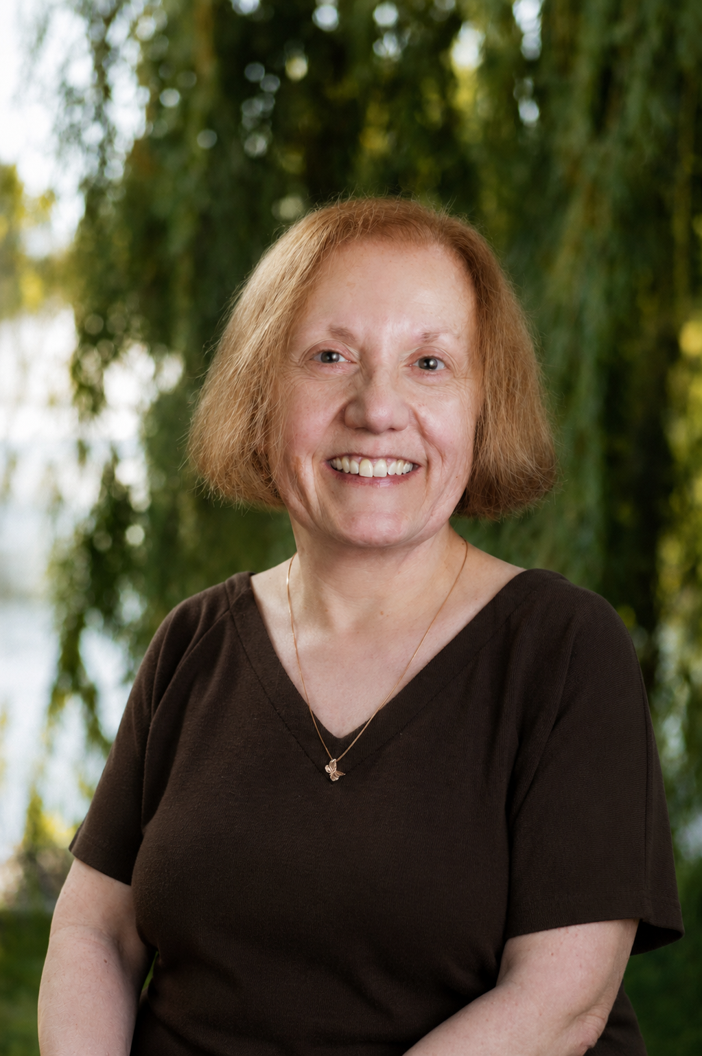 Woman with light brown hair smiles, wearing a brown shirt and a gold necklace, against a dark blue background.