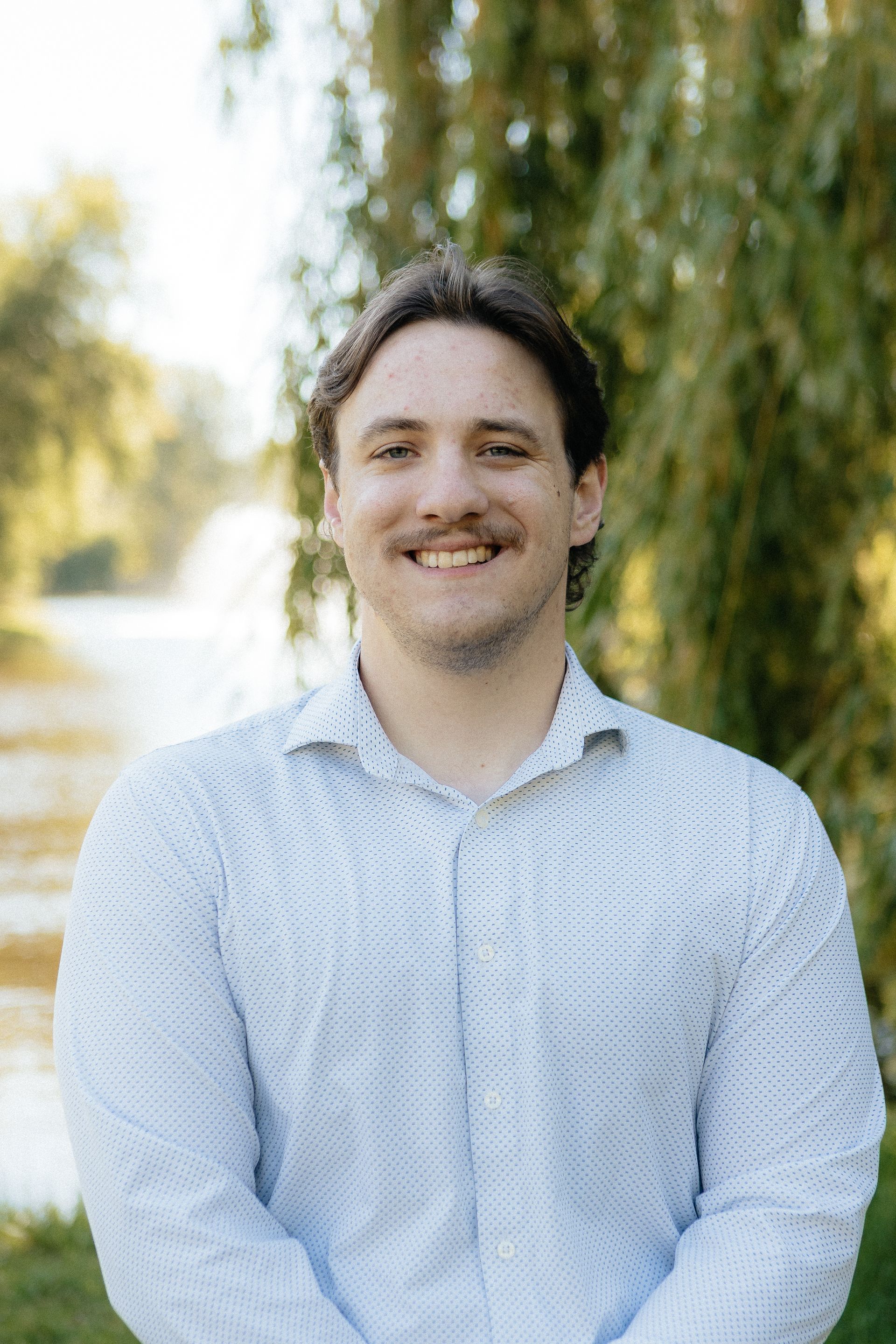 Smiling person in a light blue shirt standing outdoors with trees in the background