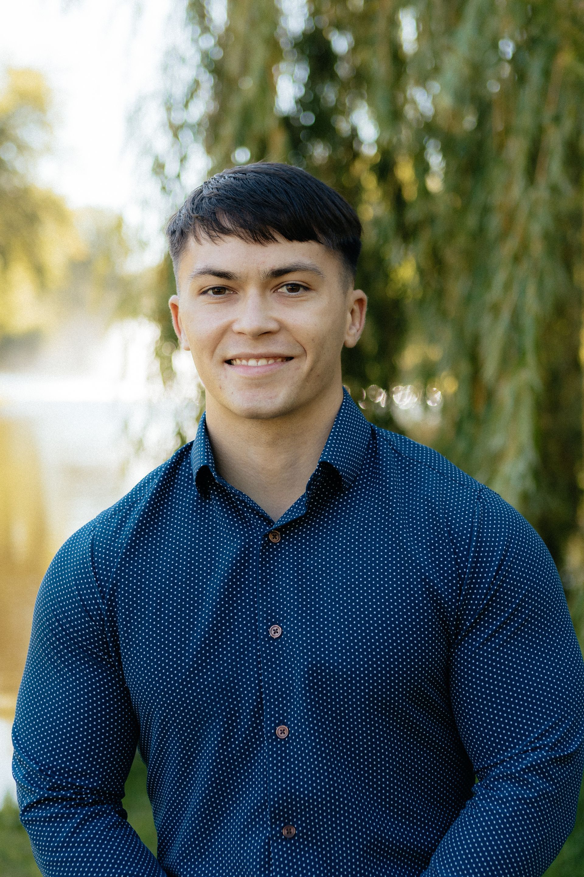 Person in a blue patterned shirt smiling outdoors with blurred trees in the background