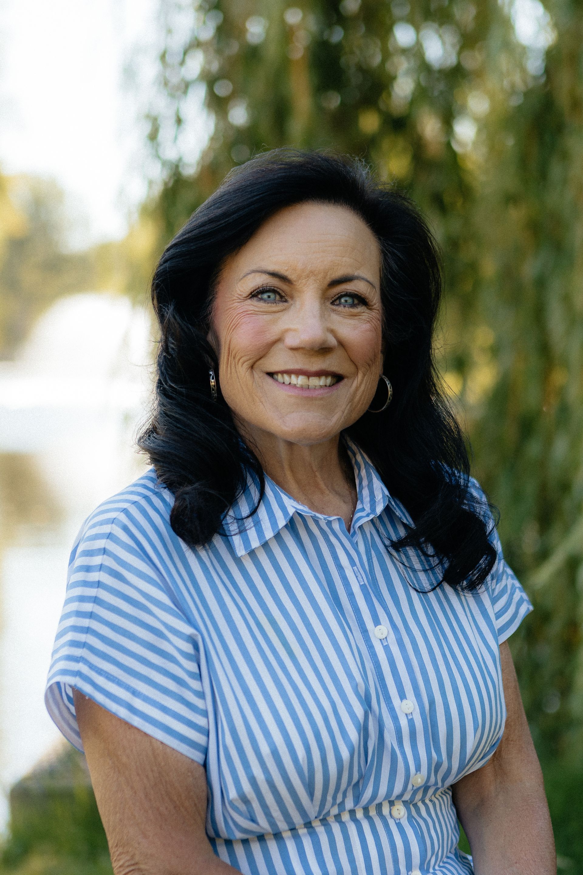 Woman with long dark hair smiles, wearing a burgundy shirt and black blazer against a blurred backdrop.
