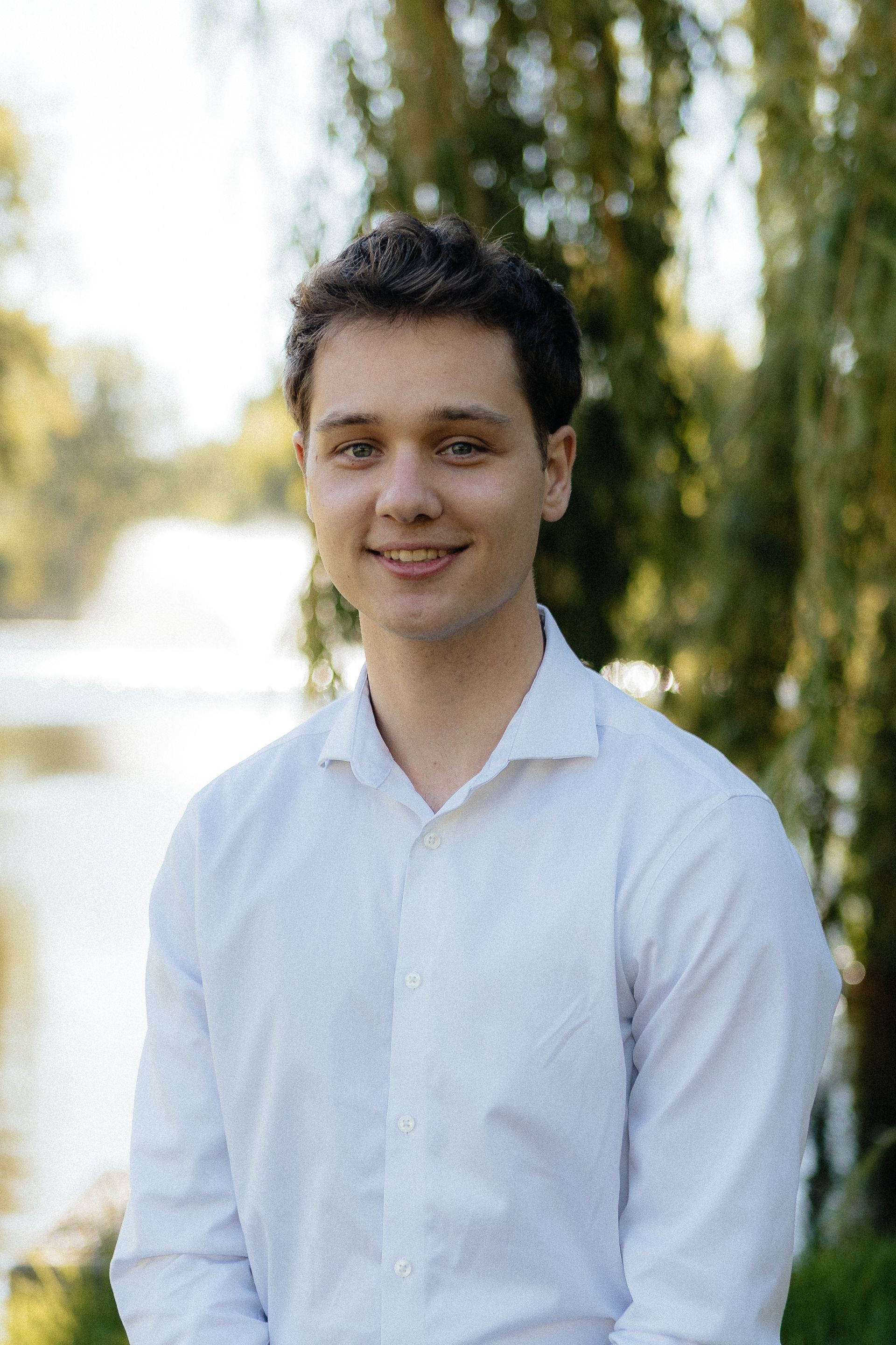 Smiling person in a white shirt standing outdoors with blurred trees and water in the background