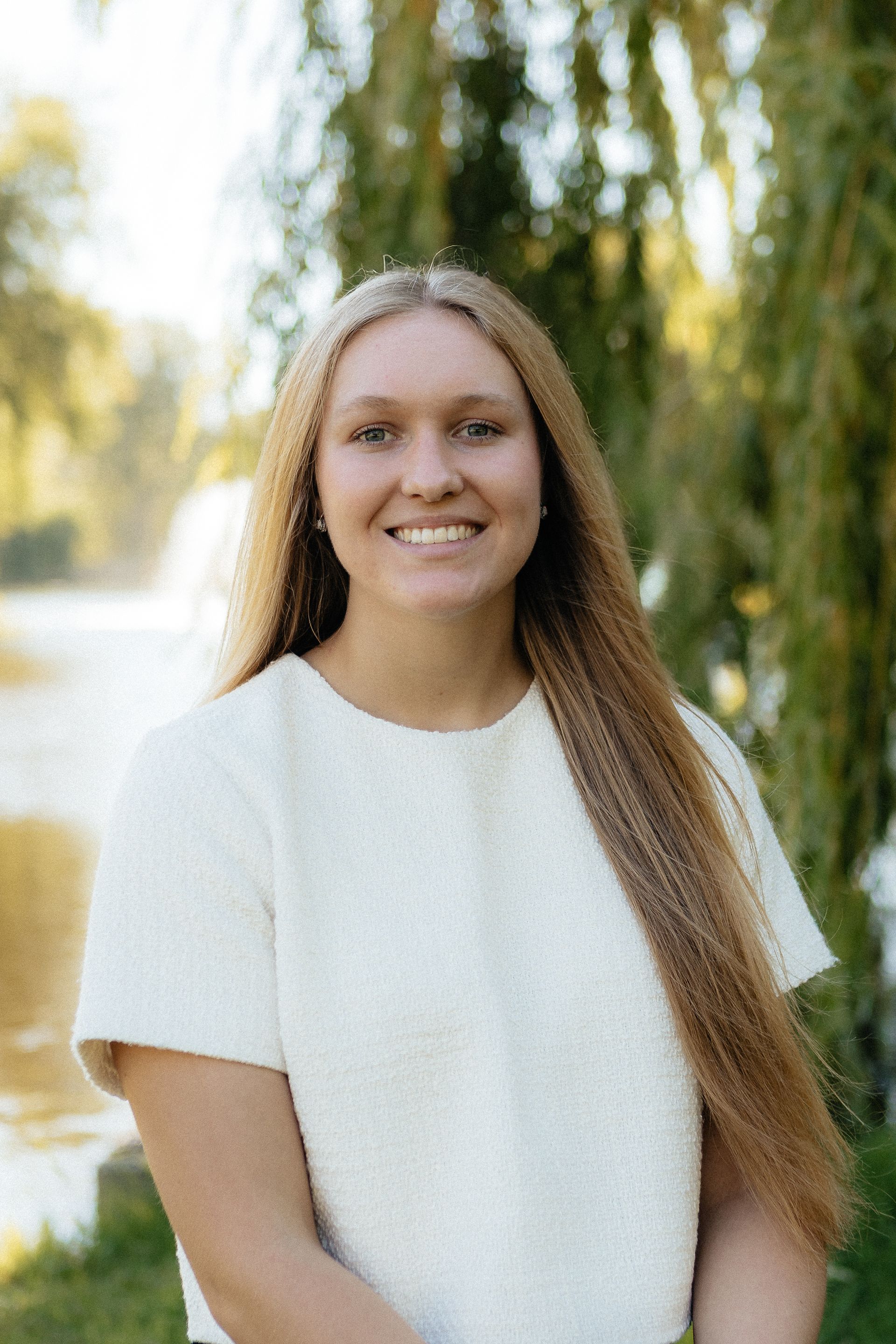 Young woman with long blonde hair smiles; white shirt, neutral background.