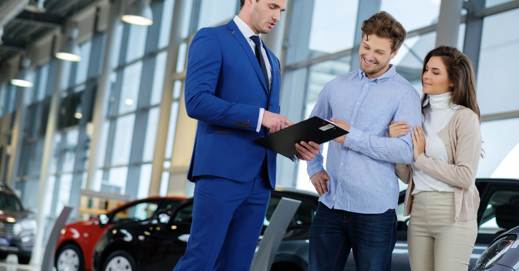 Car salesperson with clipboard talking to a couple in a car showroom; the couple is smiling.