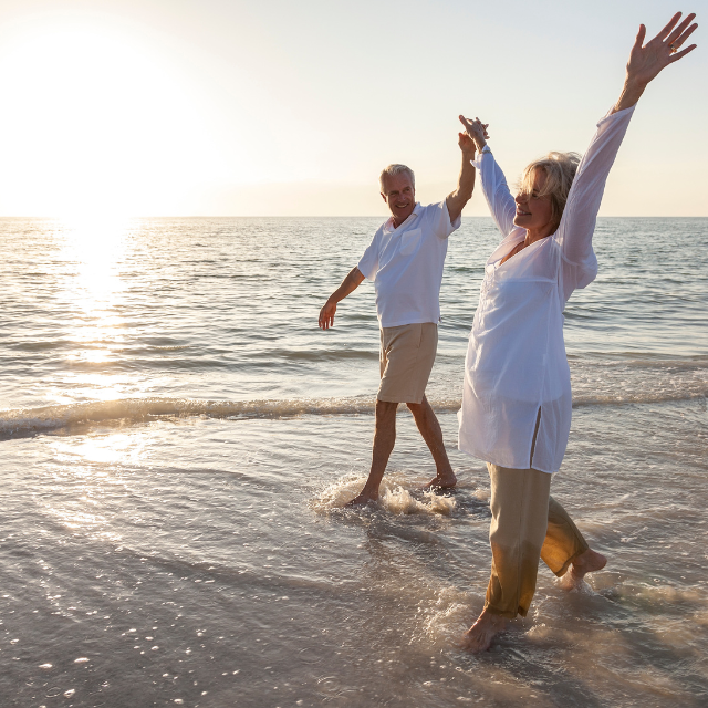 Couple at beach, holding hands, woman with arms raised, wading in water at sunset.