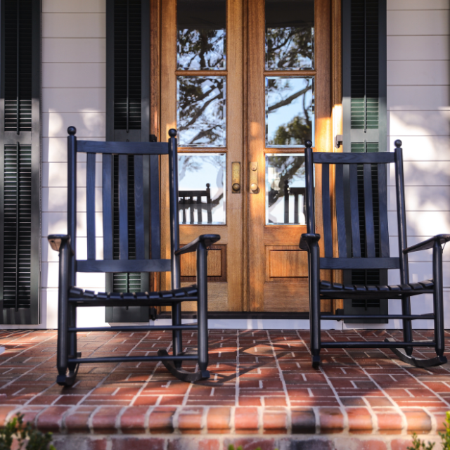 Two black rocking chairs on a red brick porch, with wooden doors and shutters in the background.