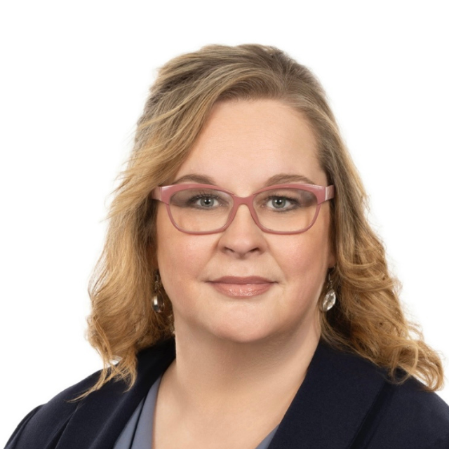 Woman with blonde hair, wearing pink glasses, a blue blazer, and earrings. Smiling, studio shot.