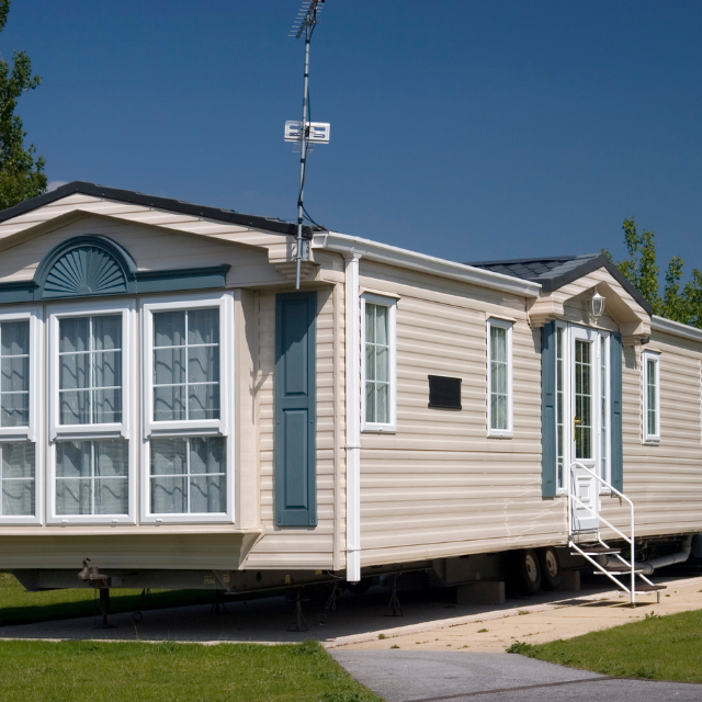 Tan mobile home with white trim and blue accents, parked on a grassy lot.