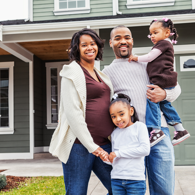 Family of four smiling in front of a green house; one parent pregnant, holding one child.