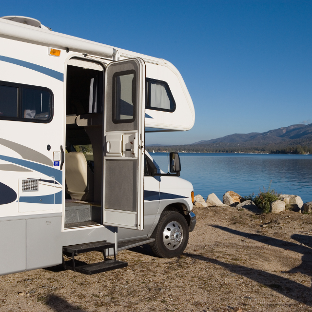 RV parked near a lake, door open, steps visible. Blue sky and mountains in the background.