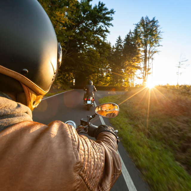 Motorcyclist wearing a helmet and leather jacket follows another motorcycle on a sunny road.