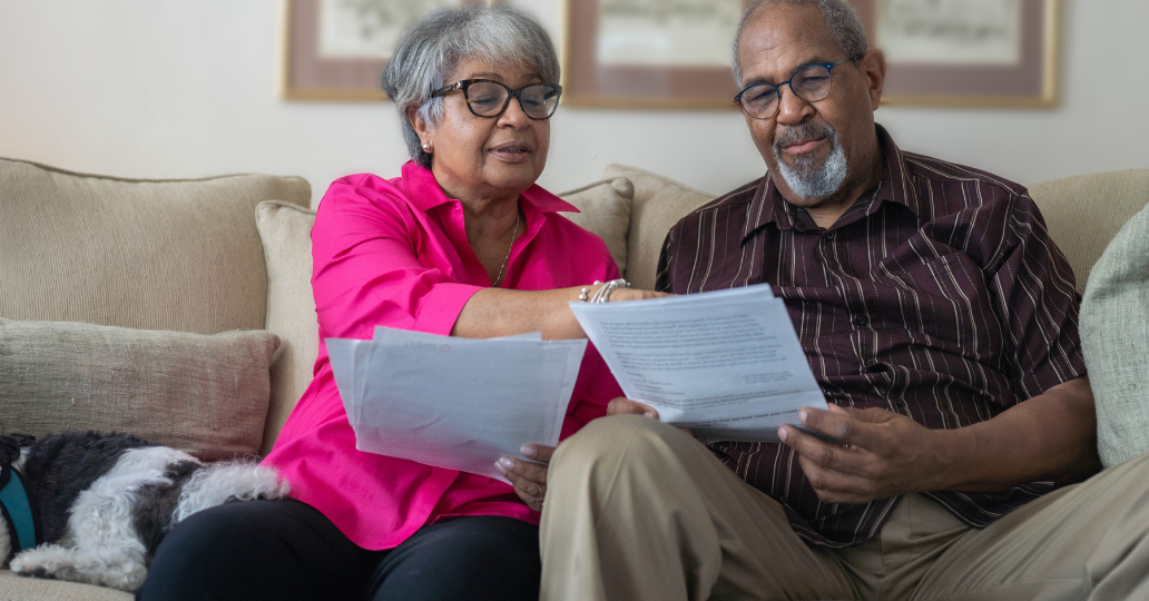 Couple reviewing papers on a couch. Woman in pink shirt points. Man in patterned shirt, glasses.